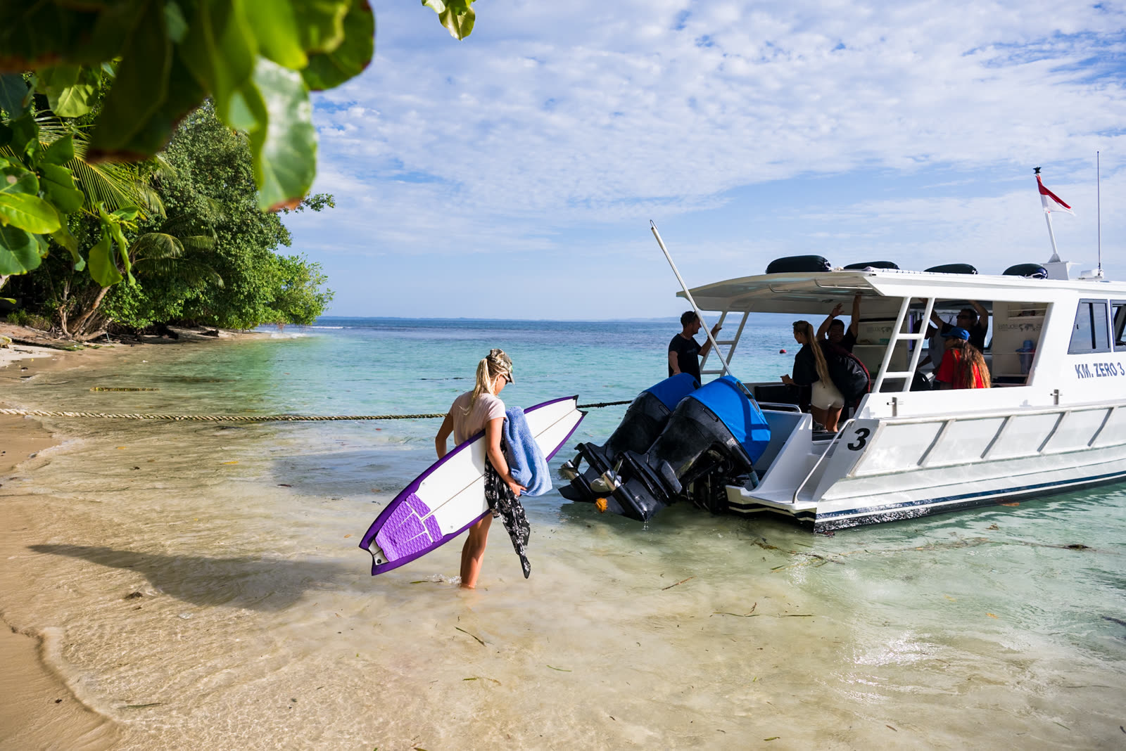 A woman wading out with her surfboard to the surf boat anchored off a jungle beach