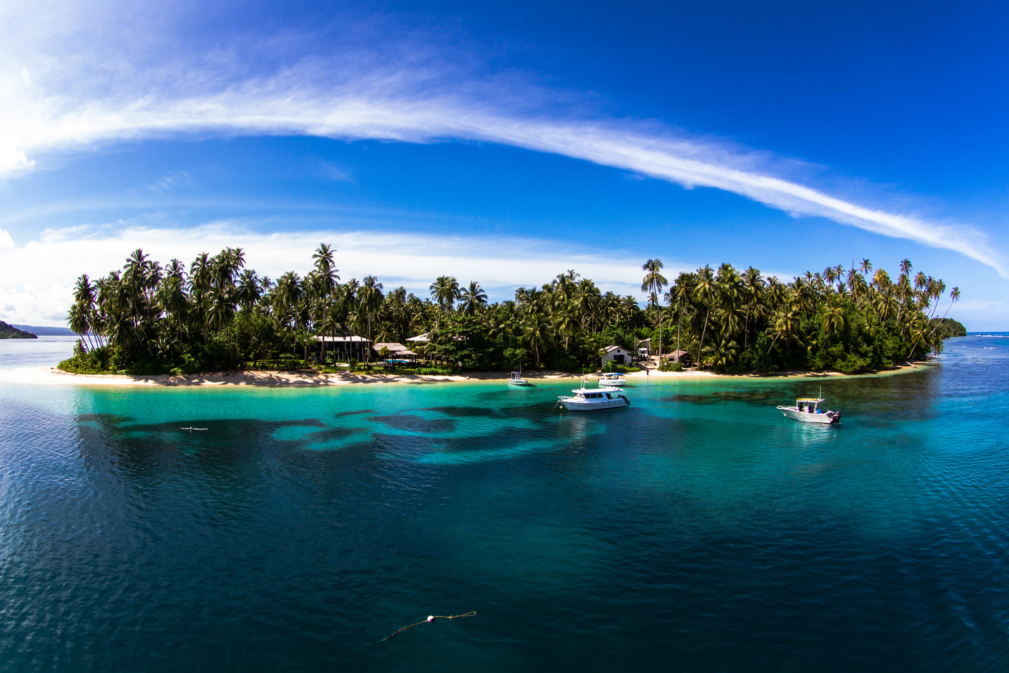 A low palm-covered island with boats anchored in clear turquoise water