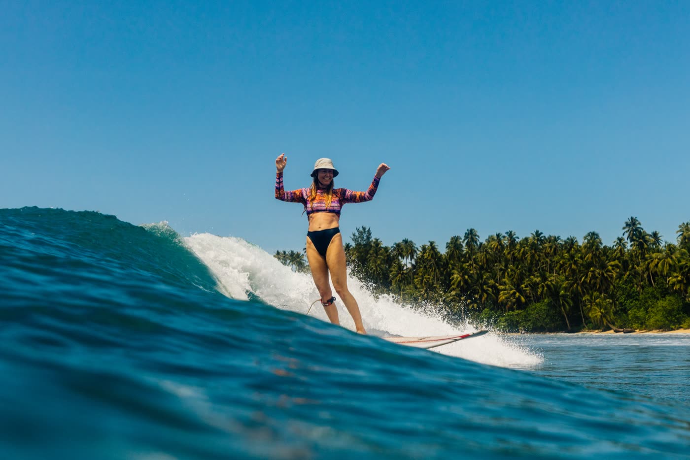 A woman on a longboard, arms out, riding a small glassy wave with palms behind