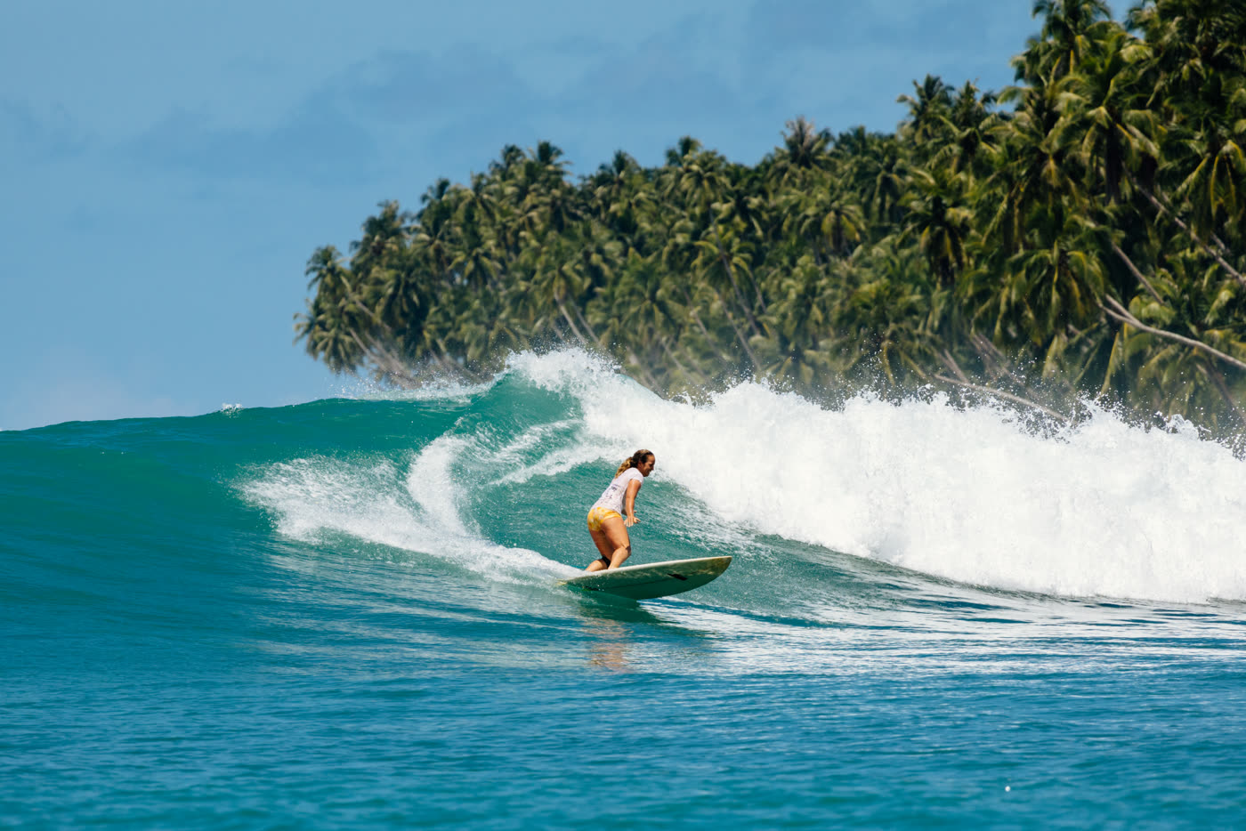 A woman on a green board racing along a bigger, peeling wave in front of palm trees