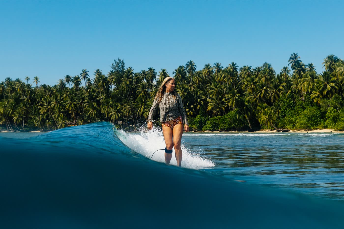 A woman in a rash vest cruising a mellow wave in front of a palm-lined island