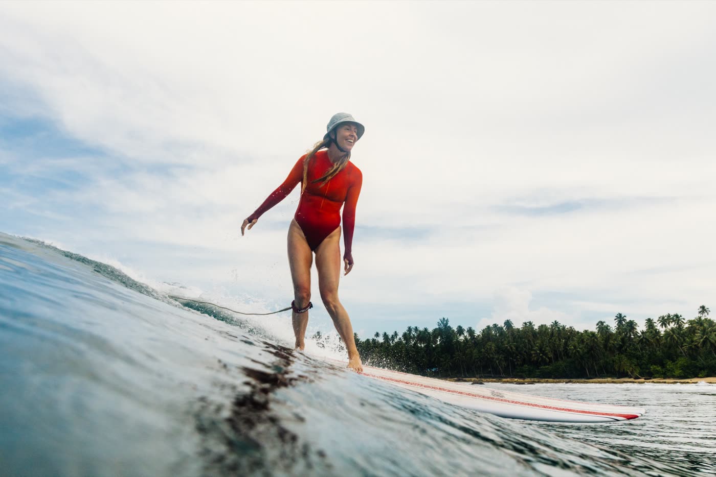 A woman in a red swimsuit and hat walking her longboard on a small wave